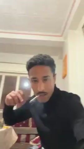 A man with short curly hair and a mustache wearing a black turtleneck sweater is standing in what appears to be a living room. He holds a cigarette in his mouth and gestures with his left fist. He then takes a drag and exhales smoke towards the camera. The room has a window, a picture on the wall, and a patterned couch in the background. There is music playing.