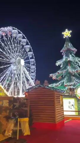The video captures a nighttime scene featuring a Ferris wheel and a large Christmas tree adorned with lights. A wooden structure resembling a small cabin sits in the foreground. The Ferris wheel is lit with white lights, and the Christmas tree is decorated with lights and tinsel. The ground appears to be covered in red material. The overall ambiance suggests a festive atmosphere, likely a Christmas market or fair.