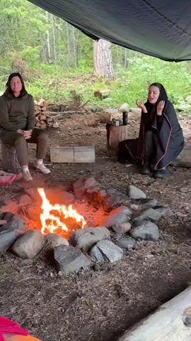 Two women are sitting around a campfire under a tarp in a wooded area. The woman on the left is wearing a green hoodie and pants and is sitting on a log. The woman on the right is wearing a black hooded garment and is sitting on the ground. A fire is burning in a ring of stones in the center. There is firewood stacked behind the woman on the left.