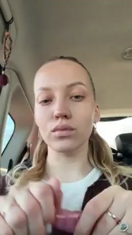 A fair-skinned woman is seated in the driver’s seat of a car. She has light eyebrows, long blonde hair pulled back in a ponytail, and is wearing simple gold earrings. Her fingernails are painted white. She wears a brown jacket over a white top. The woman opens a tube of pink lip gloss and applies it to her lips. A beaded ornament with a tassel is visible hanging from the car’s ceiling. The background includes the car’s interior, windows, and the sky outside.