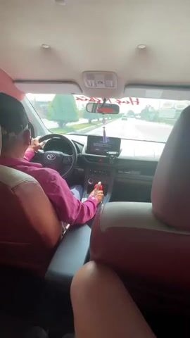 From the back seat, the camera records a taxi ride on a street. The interior of the car is a mix of red, white and black. The driver, wearing a black cap and pink shirt, can be seen adjusting his hat with his left hand. The car has a red shift knob, and a decorative item hangs from the rearview mirror. The dashboard has a screen with a purple notification icon. A partial view of a passenger's bare leg is visible at the bottom of the frame. The person recording says "Вот так вот. Сегодня меня по випу везут на такси", which translates to "That's how it is. Today I'm being driven VIP in a taxi".