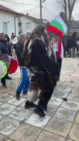 The video shows a cultural celebration taking place in an outdoor, possibly snowy setting. Several people are gathered, including some dressed in traditional costumes. There's a person in a large, shaggy, dark-colored outfit with attached bells, dancing near a man beating a green drum. Another individual is wearing a blue outfit and a tall, colorful hat, also with bells attached to their attire. People are filming the spectacle with their phones. A Bulgarian flag is visible in the background, alongside other buildings, under an overcast sky.