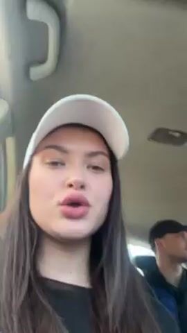 The video shows a woman in the passenger seat of a car, wearing a white baseball cap and a black shirt. She is recording herself with her phone while singing along to music playing in the car. The camera pans slightly to show a man driving, wearing sunglasses. The car appears to be moving along a highway. The woman smiles and sticks her tongue out at the camera briefly.