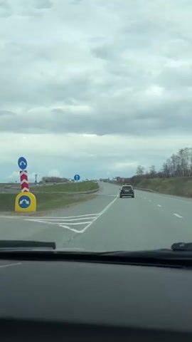 The video captures a first-person perspective from inside a moving vehicle on a multi-lane highway. The setting is a cloudy day, with a heavy, overcast sky dominating the upper portion of the frame. The road ahead is clear, with several cars visible, including a dark SUV directly in front of the camera vehicle, and other vehicles further down the highway.

To the left, an exit or junction is visible, marked by a blue circular road sign featuring a white arrow pointing diagonally right and upwards, indicating a mandatory direction. Green grass lines the sides of the highway, and sparse trees are visible on a slightly elevated embankment to the right. A metal guardrail separates the main highway lanes from the left shoulder or a parallel road. The asphalt road surface is clearly marked with white lane lines and road markings for the exit. The scene depicts typical daytime driving conditions on a highway, likely near Kulaevo, Russia, as per the provided context. No specific activities beyond driving are discernible, and no readable text is visible on any signs or vehicles.
