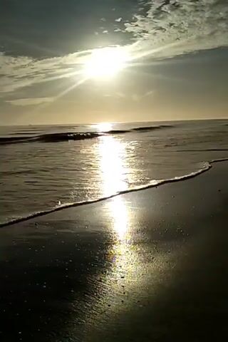 The video shows a seascape at sunrise or sunset. The sun, partially obscured by clouds, casts a bright, reflective beam across the water's surface, creating a glistening path to the shore. Small waves gently lap the sandy beach in the foreground. The sky is a mix of blues and grays with scattered clouds. The scene appears calm and serene.