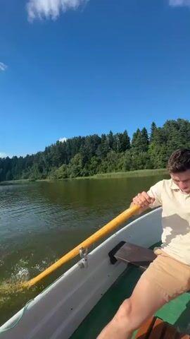 This video shows a young man rowing a boat on a lake in Lendži, Latvia. The sun is shining brightly in a clear blue sky with scattered white clouds, indicating a pleasant, sunny day. The lake is calm, with lush green trees lining the distant shore. The man is wearing a light-colored polo shirt and tan shorts, and he appears to be rowing with some effort, his face contorted in concentration. The boat is a simple rowboat, with wooden oars and a green interior. The overall scene is serene and idyllic, suggesting a leisurely activity on a beautiful summer day. There is no visible text in the video.