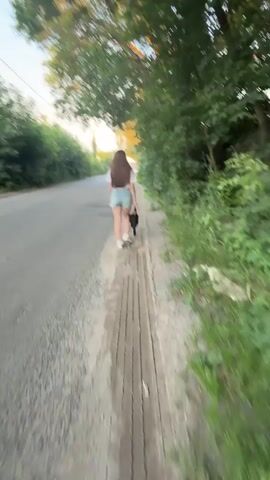The video shows a young woman walking along a paved road, with a dirt path on the right side. She is wearing a white t-shirt and denim shorts, and has long, dark hair. She is carrying a black bag in her left hand. A small black dog is walking beside her, on the dirt path.

The setting appears to be a rural or semi-rural area in Ukraine, given the presence of trees lining the road and the general atmosphere. The time of day is late afternoon or early evening, as indicated by the warm, golden light filtering through the trees. The weather seems pleasant, with clear skies and lush green vegetation.

The woman and her dog are the main subjects. The activity is a leisurely walk, possibly a stroll through the countryside. There are no vehicles immediately present in the frame with the woman, though a car is visible in the distance in one shot. The mood is peaceful and relaxed. No visible text is present in the media file.