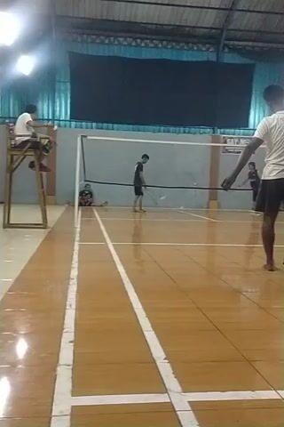 The video shows a badminton court. The court has a wooden floor with white boundary lines. A net divides the court in the middle. There are several people in the video, most of whom are men. Some are playing badminton, while others are watching or waiting. The lighting is bright, with overhead lights illuminating the court. The environment appears to be an indoor sports facility.