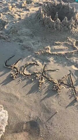 The video shows a sandy beach with a sandcastle in the background. In the foreground, the word "LUCA" is written in the sand. The tide is coming in, and waves are washing over the writing. The writing is partially submerged in water. The sandcastle is circular with crenellated walls and a hole in the center. There are also some toy buckets and spades visible behind the sandcastle. The lighting suggests it is daytime, likely late afternoon or early morning due to the angle of the shadows.