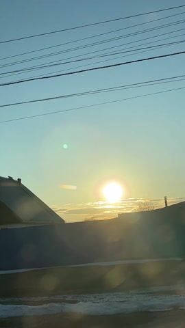 The video shows a drive-by view of a rural area. The sun is shining brightly in a clear, light blue sky. The camera is positioned in a vehicle that is moving past various buildings and residences. There are power lines visible against the sky. The houses appear to be modest, some with wood construction and traditional design elements like carved window trim. One building is identified by a sign in Russian that translates to "Hairdresser Olga". A Kia Rio is parked in front. Patches of snow remain on the ground.