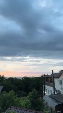 This is a vertical video clip captured from a high vantage point, likely a balcony or window, overlooking a wooded area with several houses. The scene is set during a sunset, as indicated by the soft, warm hues of pink and orange painting the sky near the horizon. Dark, heavy clouds dominate the upper portion of the sky, suggesting a dramatic or possibly approaching rain, but the light filtering through them creates a beautiful, serene contrast.

The subjects are primarily the natural landscape and man-made structures. Lush green trees form a dense canopy, partially obscuring the houses below. The houses appear to be rustic, with visible wooden log construction and dark-colored roofs. One house in the foreground has a distinct green roof and several windows. The overall impression of the setting is one of a secluded, natural environment, perhaps a countryside retreat or a village nestled amongst the trees.

No people are visible in the frame, and the activity is static, focusing on the visual experience of the sunset. Notable details include the texture of the clouds, the varying shades of green in the foliage, and the architectural style of the houses. The time of day is clearly evening, given the sunset colors. The weather appears to be a transition, with both stormy-looking clouds and the gentle light of sunset present. The location cues, such as the style of the houses and the dense forest, combined with the provided context of Voronov, Russia, suggest a rural or semi-rural setting in that region. The emotion evoked is one of tranquility and natural beauty, with a hint of dramatic atmosphere due to the clouds. There is no visible text in the media.