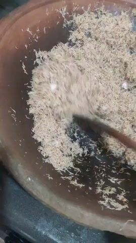 A close-up shot shows shredded coconut being stirred in a clay pot. A wooden spoon is used to move the coconut, which appears to be toasting. The pot is round and has a rustic, earthy appearance. The coconut is light brown and dry, suggesting it is being prepared for a dish. The background is blurred but appears to be a kitchen environment. The lighting is natural and soft, creating a warm and inviting atmosphere.
