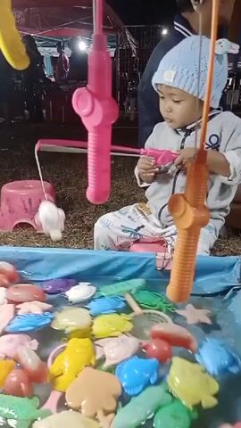 A young child wearing a blue knitted hat and a grey sweatshirt with white patterns is playing a fishing game. The child is sitting in front of a blue plastic tub filled with water and various colorful plastic sea creatures, including fish, turtles, and crabs. The child is holding a pink fishing rod with a magnetic hook, attempting to catch the floating toys. Two other fishing rods, one pink and one orange, are visible hanging above the tub. In the background, blurry figures and stalls suggest an outdoor market or fair. The ground appears to be covered in grass and dirt. The lighting indicates it is nighttime.