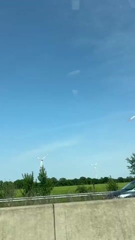 The video shows a view of several wind turbines from a moving vehicle. The turbines are white with red accents on the tips of the blades. They are located in a field of green grass with some trees in the background, under a bright blue sky with sparse clouds. A concrete barrier is visible in the foreground, separating the road from the field. There are other vehicles passing on the road.