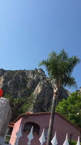 The video begins by showing a pink building with white flags in the foreground, a palm tree, and a rocky mountain in the background under a clear blue sky. The camera then pans to show lush green foliage and red flowers against the sky. Next, the video shows a street scene with people walking on a sidewalk next to a road. A man in a blue hat and patterned shirt walks away from the camera. Pink flowers are visible on a bush to the left of the frame. A rocky cliff with buildings on top is visible in the background, as is a light pole and a car. The video ends showing the same street scene with the cliff and buildings in the background.
