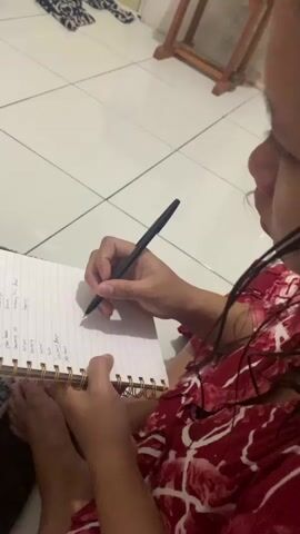 A person is shown from the chest down, sitting and writing in a spiral-bound notebook with a black pen. The person is wearing a red and white patterned garment. The notebook is open to a page with handwritten text. The person's bare feet are visible, resting on a patterned rug. The floor is tiled, and the background shows a wall, a piece of furniture, and kitchen elements including shelves and containers. The lighting appears to be artificial.