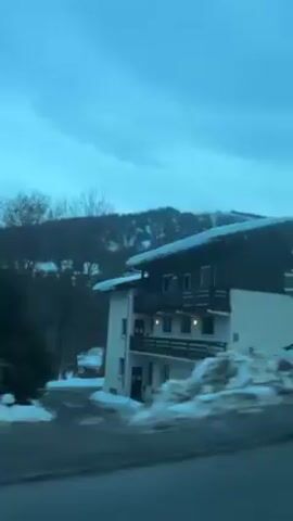 The video shows a moving perspective of a snowy landscape with houses, taken from a vehicle. The sky is a muted blue with scattered clouds. The houses are a mix of dark wood and white structures, some with snow-covered roofs. A snow-covered mountain is visible in the background. Cars are parked near the houses, and a street runs in front of them. The camera moves past the scene.