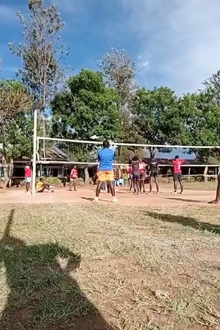 The video shows a group of people on a volleyball court. Some are stretching near the net, while others stand around. A person in a blue shirt and orange shorts stands on the court facing the net, seemingly in a ready position. The volleyball net is strung between two white poles. Trees and a low building are visible in the background under a blue, slightly cloudy sky. The camera's view is partially obstructed by grass and a large shadow in the foreground.