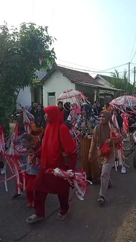 A group of children and adults are walking down a paved road, possibly in a procession. The children are mostly wearing red and gray uniforms. Several adults are wearing headscarves and traditional Indonesian clothing, including batik. Some individuals are carrying decorative items, such as umbrellas adorned with red and white streamers. The environment appears to be a rural or suburban area, with houses and vegetation visible in the background. The sky is bright, suggesting daylight.