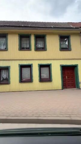 This is a video of a yellow house with green trim. There are several windows on the front of the house, all with dark red frames. The front door is also red. The house is made of wood siding. In front of the house is a brick walkway. The sky is overcast.