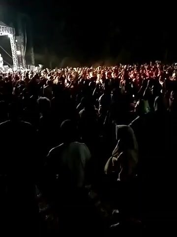 A nighttime outdoor concert is in progress. A large crowd of people is gathered in front of a raised stage where a band is performing. The stage is lit with bright white and colored lights, casting a glow on the performers and the front rows of the audience. The rest of the crowd is largely in shadow, with only faint outlines visible. The overall atmosphere appears to be energetic and lively, with the audience seemingly engaged with the performance.
