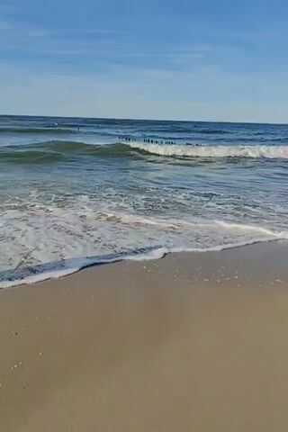 The video shows a beach with sand in the foreground and waves gently rolling onto the shore. The water is a clear blue-green, and the sky above is a bright blue with a few wispy clouds. In the distance, there is a line of wooden posts partially submerged in the water, likely a breakwater. The scene is serene and peaceful, with the sound of the ocean waves providing a natural soundtrack. There are no people or animals visible in the video. The lighting suggests it is daytime, likely morning or afternoon.