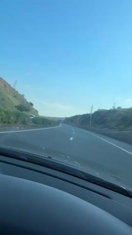 The video shows a point-of-view shot from inside a vehicle, looking out through the windshield at a highway. The road has multiple lanes and is flanked by hills on both sides, covered in sparse vegetation. The sky is clear and blue. Power lines are visible along the sides of the road. The vehicle is in motion, as indicated by the blurred lines on the road.