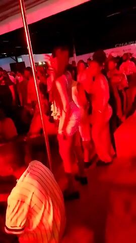 The video shows a group of people dancing at a party. The room is lit with red lights, and there is a dance pole in the middle of the floor. One woman in a sparkly dress is dancing near the pole, while others move freely around the room. Some of the partygoers are dressed in animal print. The room is crowded, and the atmosphere is festive.