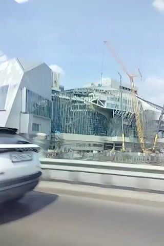 A white SUV is driving past a large building under construction. The building appears to be a modern structure with a lot of glass and angular shapes. There are cranes visible at the construction site. The sky is blue with some clouds. The video is shot from inside the SUV, looking out the passenger window.