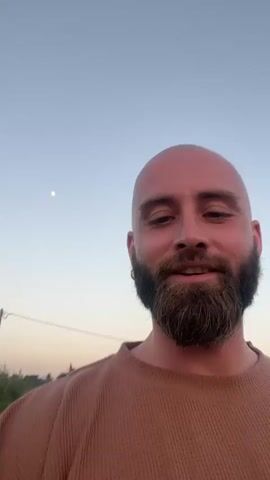 A bald man with a beard is filming a selfie video on a concrete walkway in front of a rice paddy landscape. Behind him are five younger boys, who are waving, smiling, and shouting at the camera. The sky is light blue, suggesting either early morning or late afternoon.