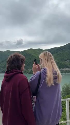 Two women stand on a balcony overlooking a large lake and mountains. The woman on the left wears a maroon hoodie and the woman on the right wears a lilac hoodie and a pink fanny pack with a chain. The woman on the right holds up her phone and takes a photo of the woman on the left. The lake is a light blue color and is surrounded by green mountains. The sky is overcast with gray clouds.