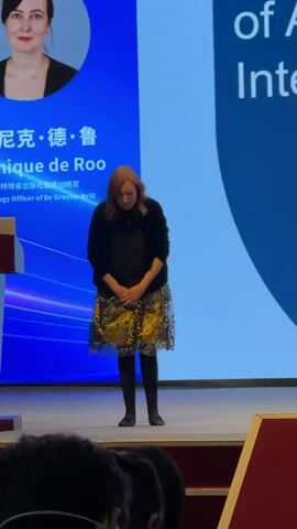 The video shows a woman standing on a stage, in front of a podium and a large screen. The screen displays text in both Chinese and English, with a portrait of the woman at the top left. The text on the screen identifies her as Dominique de Roo, Chief Strategy Officer of De Gruyter Brill. The podium has an emblem with Chinese characters and the words "INSTITUTE OF GRAPHIC COMMUNICATION 1958". The woman is wearing a black top, a patterned skirt, and dark tights. She is initially bowing, then stands up and walks to the podium, where she takes the microphone and appears to begin speaking. There are people in the foreground, whose heads are visible, suggesting the audience is seated. The lighting is bright, likely from stage lights. The overall environment appears to be a formal presentation or conference setting.