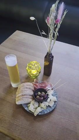 This media file captures a close-up, top-down view of a table setting in what appears to be a restaurant or bar. The primary subjects are a glass of light-colored beer with a foamy head, a small appetizer plate, a decorative candle, and a vase with dried flowers.

The appetizer plate is round and appears to be made of ceramic with a textured rim. It contains a selection of food items including slices of bread, cured meat (likely prosciutto or ham), cubed cheese, olives, and what look like artichoke hearts. Several thin wooden skewers are also present on the plate, suggesting that some of the food items are intended to be picked up with them.

A tall, clear glass filled with beer sits to the left of the plate. To the right of the beer and behind the plate is a small, ornate candle holder with a textured, green glass exterior, containing a lit candle that emits a warm glow. Behind the candle holder is a dark brown glass vase holding a small arrangement of dried grasses and a few pink and white feathery flowers.

The table surface is made of wood with a visible grain pattern. In the background, a dark-colored upholstered bench or seating area is visible, suggesting an indoor setting. The lighting appears to be dim and ambient, possibly from the candle and overhead restaurant lighting, giving the scene a cozy and intimate feel. No people are visible in the frame. There is no visible text in the media file. The overall impression is that of a relaxed moment, perhaps a pre-meal aperitif or a light snack. The time of day cannot be definitively determined, but the dim lighting suggests it could be evening.