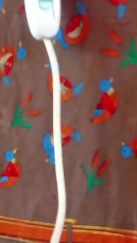 The video shows a close-up of a white cord hanging down against a brown fabric background decorated with colorful cartoon birds and cattails. The cord originates from a white object at the top of the frame, which appears to be a power adapter or a similar electronic device. The cord is long and slightly curved, and it hangs vertically in the center of the frame. The background fabric has a repeating pattern of birds in various colors, including red, blue, and orange, and some green cattail plants. The lighting is bright, illuminating the cord and the background clearly.