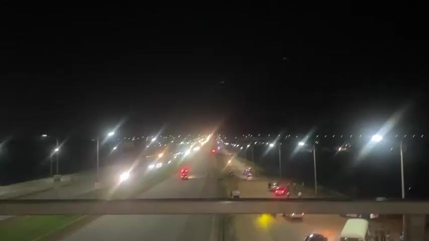 This media file captures a nighttime scene on a road in Sauka, Nigeria. The primary subject is a multi-lane road illuminated by streetlights, with vehicles moving along its lanes. The setting appears to be a bridge or elevated roadway, as suggested by the perspective and the presence of a railing in the foreground. 

Several cars and a bus are visible on the road, their headlights and taillights creating streaks of light due to the camera's movement or a long exposure. On the right side of the road, a gathered crowd of people are visible, some standing and others near parked vehicles or makeshift stalls. The atmosphere suggests a busy or perhaps a festive gathering, with the presence of numerous people and vehicles. 

The time of day is clearly night, with a dark sky and artificial lighting dominating the scene. There are no explicit weather cues, but the darkness and clear visibility of lights suggest it's likely a clear night. The location is identified as Sauka, Nigeria. No discernible text is visible in the media file. The overall impression is one of a lively night scene, with traffic and a public gathering taking place.