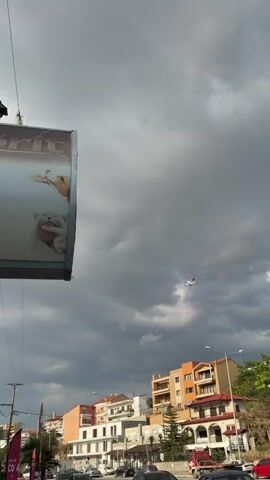 A small white plane with red accents flies through a cloudy sky. The plane is visible in the center of the frame against a backdrop of dark, dramatic clouds. In the foreground, on the left side of the image, a portion of a billboard is visible. The billboard appears to feature an image of a cat. Below the plane and in the distance, the tops of buildings with orange and beige facades are visible. The overall impression is of an overcast day with some interesting aerial activity.