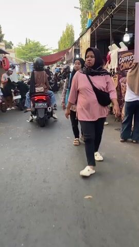 The video shows a street scene, possibly a market or a busy road with stalls. Several people are walking or riding on motorcycles. In the foreground, a woman wearing a pink shirt and black pants with a black fanny pack walks towards the right. To her left, another woman wearing a black hijab and a plaid shirt walks in the same direction. Behind them, a motorcycle with two people on it drives away from the camera. Further in the background, there are more people and motorcycles, as well as stalls with merchandise. The overall atmosphere appears to be daytime, with a slightly overcast sky.