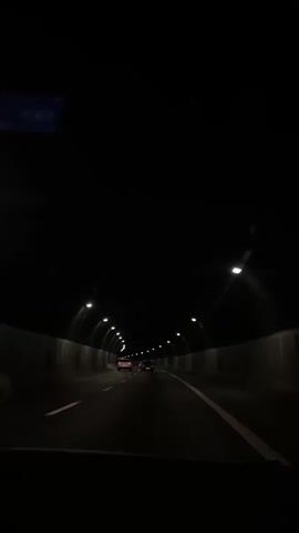 The video shows a view from inside a vehicle driving through a dark tunnel. The tunnel is illuminated by regular intervals of bright, circular lights lining the ceiling. The walls of the tunnel are visible on either side, appearing to be made of concrete or stone. Several other vehicles are visible ahead, their taillights creating streaks of red light in the darkness. The road surface is dark and appears to be asphalt, with white lane markings visible. The perspective suggests the camera is positioned behind the windshield of the vehicle.