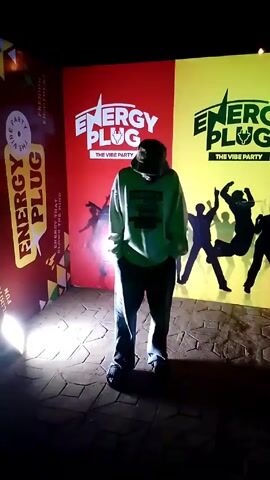 The video shows a person standing in front of a backdrop promoting "Energy Plug, The Vibe Party". The backdrop consists of three panels: the left panel is mostly red, the middle panel is red, and the right panel is yellow. The person is wearing a light green sweatshirt with the word "GEOMATRICAL" printed on it, dark jeans, black sandals, and a dark bucket hat. The person initially stands with their hands in their pockets, then makes a peace sign with their left hand. The flooring is made of light grey paving stones. The lighting is somewhat bright, creating shadows on the backdrop. Musical audio is also present.