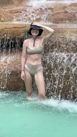 A young woman stands in a pool of light-blue water, in front of a small artificial waterfall. She wears a khaki-colored bikini and a dark-colored bucket hat. Her right hand is on her head as if adjusting the hat, while her left arm hangs at her side. The waterfall cascades down a rocky structure behind her. The water is turbulent around her legs.