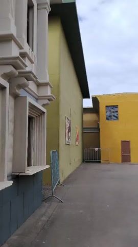 The video pans across an outdoor courtyard area with several buildings. On the left, a light-colored, ornate building facade with windows is visible. Adjacent to it is a pale yellow building with posters on the wall and a blue metal barrier in front. Further into the courtyard, a small yellow building with a brown door and a single window is seen, with a metal fence in front of it. In the background, a large, gray building with arched entrances and decorative detailing stands out. To the right of that, there are red and pink structures, including what appear to be small, wooden guard booths. The video continues to pan, showing a multi-story pink building with numerous windows and awnings, as well as dark storefronts on the ground level. The ground is paved with asphalt, and the sky is overcast with clouds.