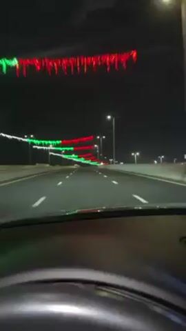 The video shows a car driving on a highway at night. The road is illuminated by streetlights and decorated with strings of red, green, and white lights strung between the poles. The dashboard and steering wheel of the car are visible in the foreground. There is minimal traffic.