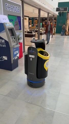 A gray and black self-driving trash can with yellow accents and the words "ATALIAN GLOBAL SERVICES" on it stands in a tiled shopping mall, illuminated by blue lights at its base. Behind it, an older man in a brown jacket and jeans walks towards the camera. To the left of the trash can is an ATM. Beyond the man, other people walk in the distance in front of shops.