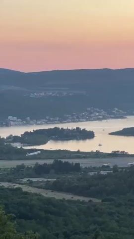 This is a vertical, long shot video of an airport runway near a body of water, with rolling hills and a town in the background. The sky displays a soft gradient of pink and orange, indicating either sunrise or sunset. An airplane is captured in mid-flight, just having taken off from the runway. The plane is white with red accents on its tail. Several sailboats are visible on the water. The foreground is dominated by dark green trees.