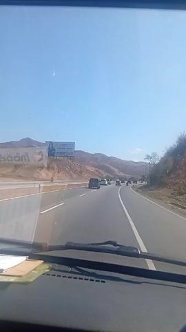 The video is taken from the inside of a moving vehicle on a highway during the day. There are several other vehicles on the road, traveling in the same direction. In the distance, mountains can be seen against a clear blue sky. A large billboard is visible on a hillside. The dashboard and windshield wipers of the vehicle recording the video are visible.