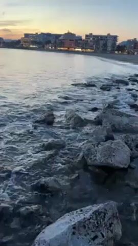 The video shows a coastal scene at dusk. Waves gently lap against a rocky shoreline in the foreground. Beyond the rocks is a sandy beach. Further in the background, buildings are illuminated, suggesting a small town or resort area. The sky transitions from a pale blue to an orange hue. The overall scene is calm and peaceful.