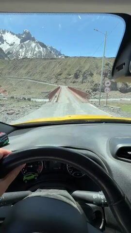 The video shows the interior view of a yellow vehicle driving towards a narrow bridge in a mountainous area. The snow-capped mountains are visible in the background under a bright blue sky. The bridge has red railings and a weathered concrete surface. The car's dashboard is visible, including the steering wheel, speedometer, and other gauges. A hand is visible on the steering wheel. There are some small items on the dashboard as well. The car appears to be the only vehicle on the bridge at the moment.
