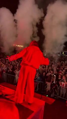 A person in a red tracksuit stands on a platform on a stage. Smoke is rising in the background and pyrotechnics are visible. A large crowd of people is gathered in front of the stage behind a barrier. The scene appears to be an outdoor concert or festival at night. The person on stage is facing away from the camera, towards the crowd.