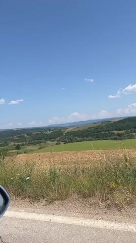 This is a landscape video, shot from a moving vehicle, showing a rural scene under a clear blue sky with scattered clouds. The foreground features a patch of dry grass and weeds along the side of a road. Beyond this, a golden field of harvested crops or dry grass stretches across a gentle slope. Further in the distance, there are rolling green hills covered in trees and fields, with a few visible structures, possibly farms or rural dwellings. The overall impression is one of a sunny, rural summer day.