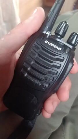 A hand holds a black Baofeng walkie-talkie. The walkie-talkie has a black antenna and two black knobs on top. The word "BAOFENG" is visible in white lettering on the front of the walkie-talkie. The background is out of focus but appears to be indoors with some wooden elements and fabric.